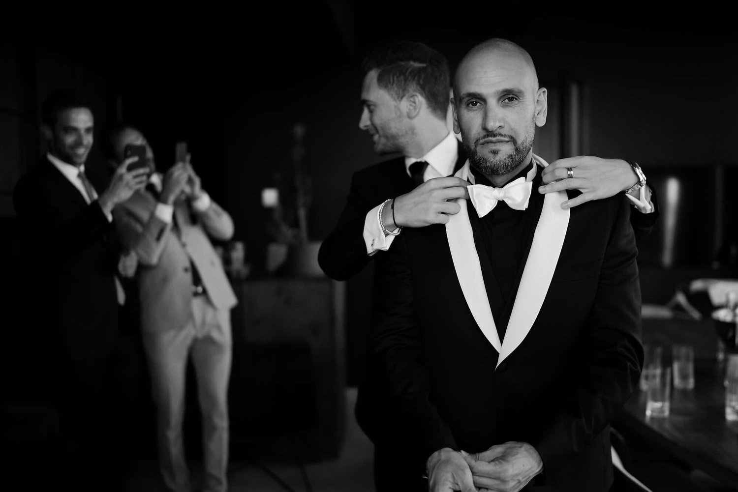 Kareline_Amir_021_2 - Best man adjusting groom bow tie while guests taking photos.jpg